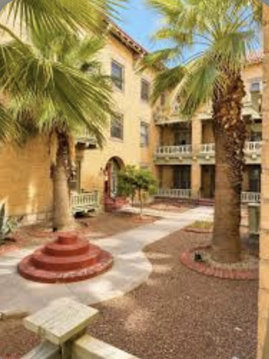 Palm Court courtyard with palm trees, arched balconies and entrances, red brick planters, and blue sky